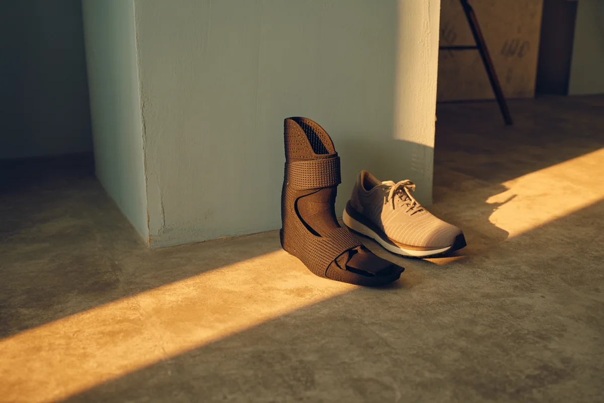 A moulded ankle-foot orthosis brace propped against a pale plastered wall beside a single walking shoe, with a long raking shadow in golden-hour light