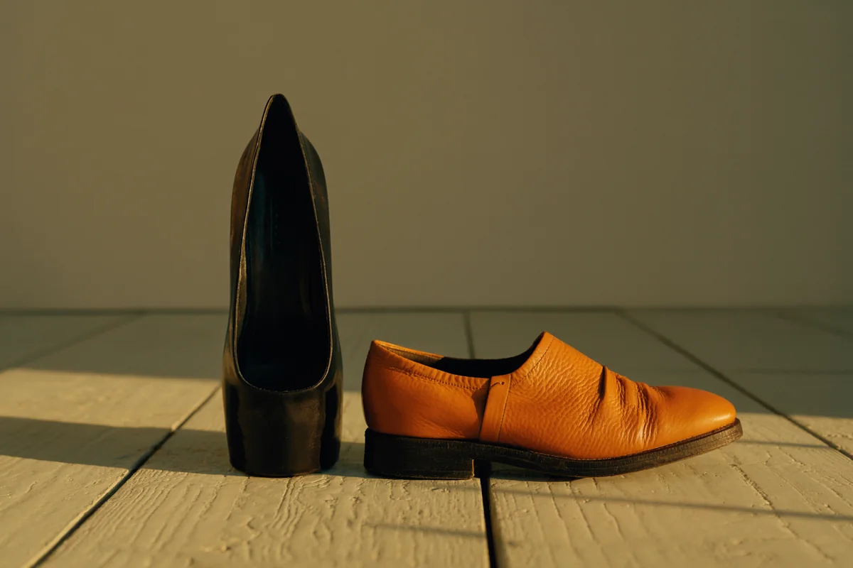 A narrow pointed leather dress shoe standing beside a broad round-toed leather walking shoe on pale weathered floorboards in golden-hour light