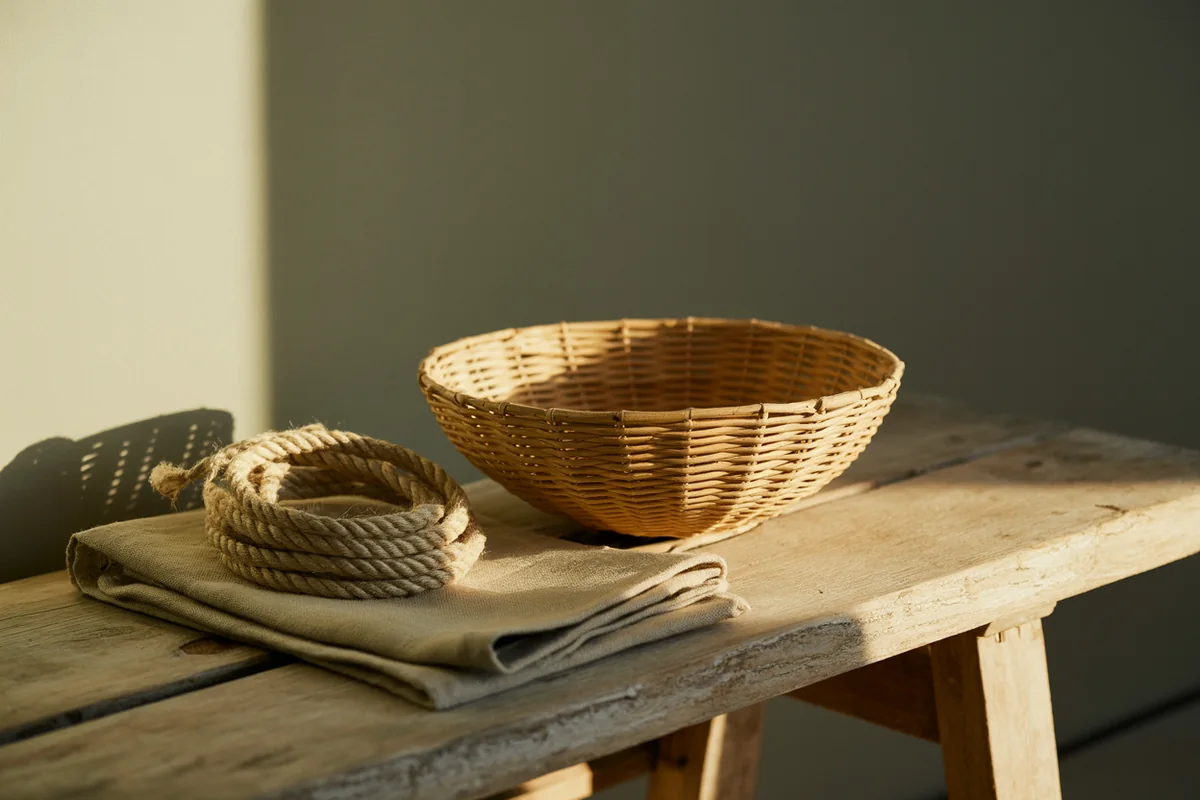 A handwoven rattan bowl resting on its side beside jute rope and sage linen on a weathered oak bench in golden-hour light