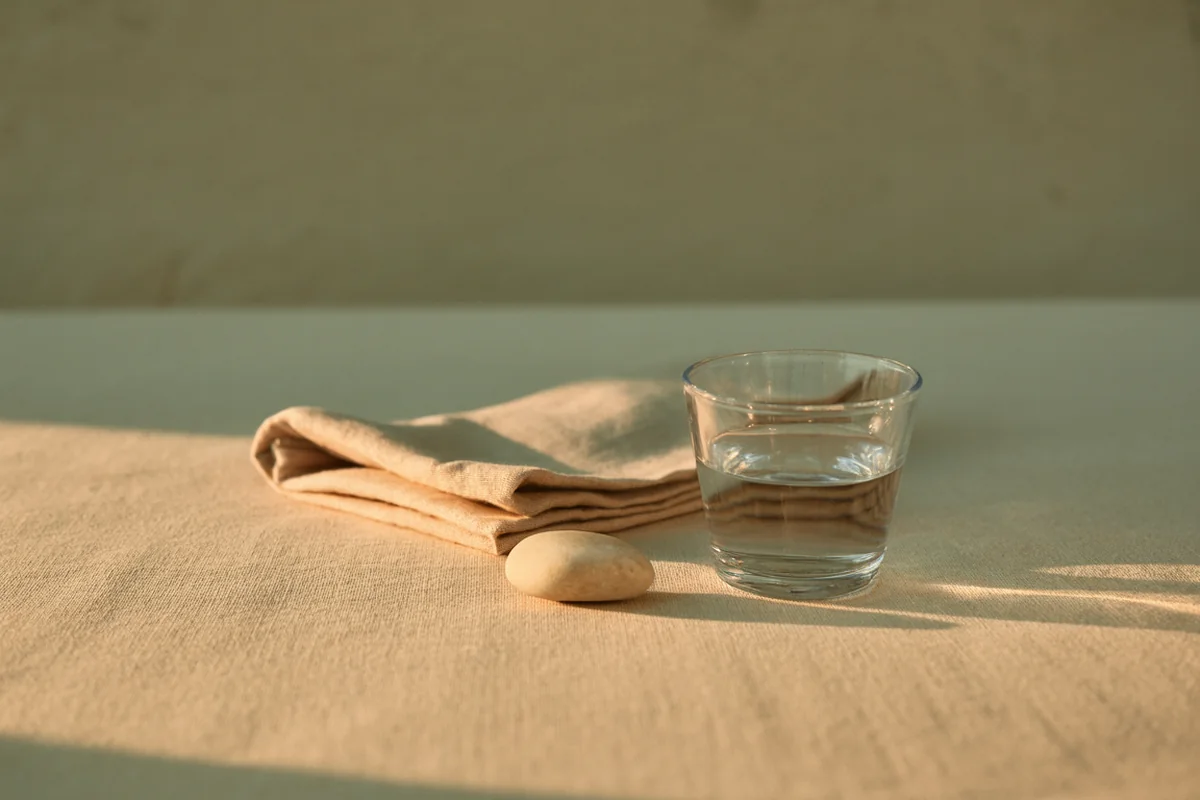 A single clear glass vessel of clear liquid beside folded linen and one pale pebble in warm afternoon light